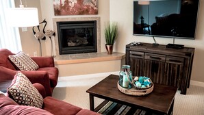 Una sala de estar con chimenea, televisor, una mesa de centro con objetos decorativos y un sofá rojo.