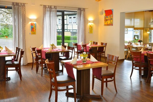 A restaurant with wooden tables and chairs, large windows with curtains, and artwork on the walls. - Hotel Restaurant Zum Mühlental (Buch)