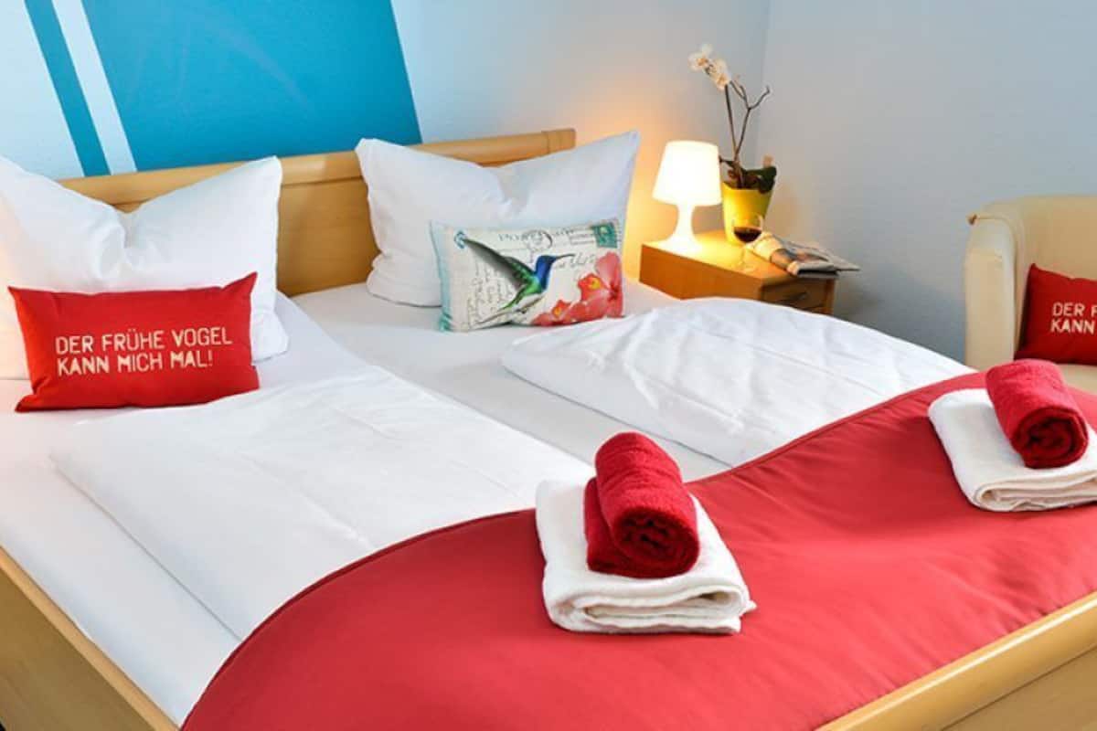 Ein Hotelzimmer mit zwei Betten, rot-weißer Bettwäsche und Kissen mit Aufschrift. An den Betten stehen Nachttische mit Lampen, und ein Beistelltisch trägt eine Lampe sowie eine Vase mit Blumen.