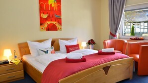 Ein Hotelzimmer mit einem hölzernen Bett, roter Bettwäsche, einem Nachttisch mit Lampe, einem Fenster mit Vorhängen und einem Gemälde an der Wand.