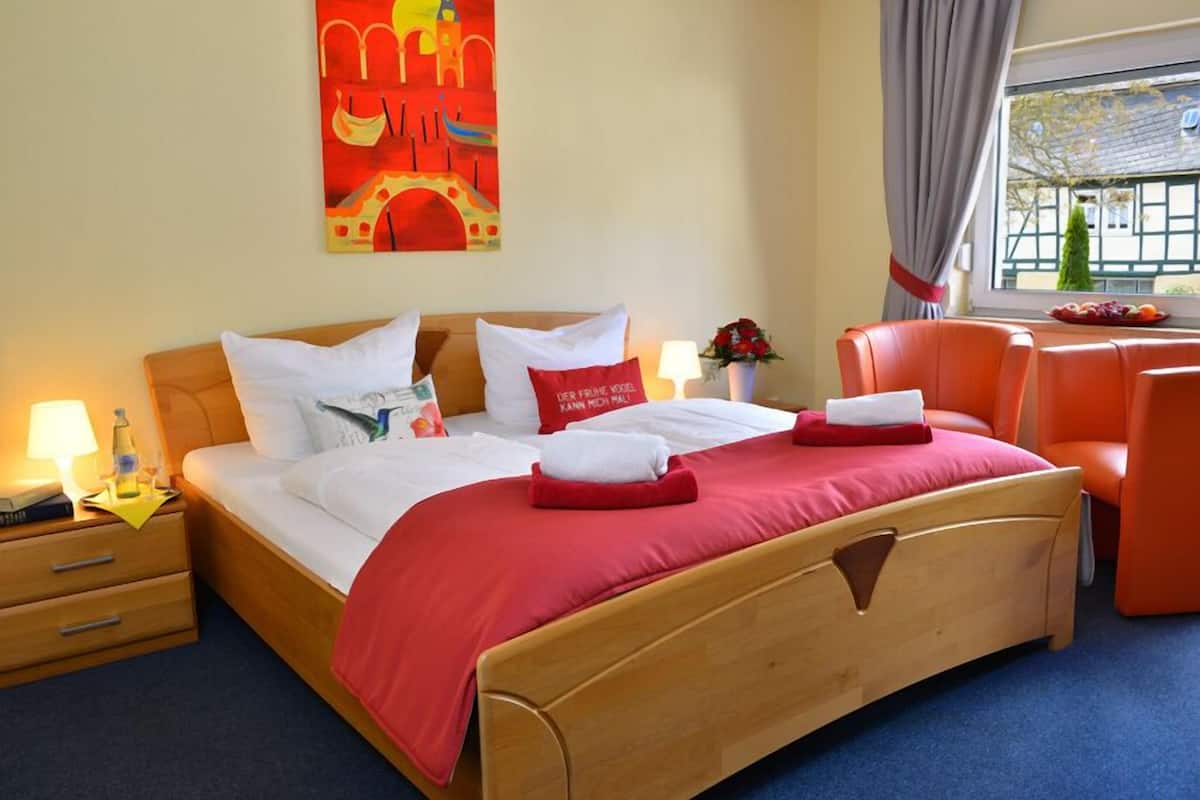 Ein Hotelzimmer mit einem hölzernen Bett, roter Bettwäsche, einem Nachttisch mit Lampe, einem Fenster mit Vorhängen und einem Gemälde an der Wand.