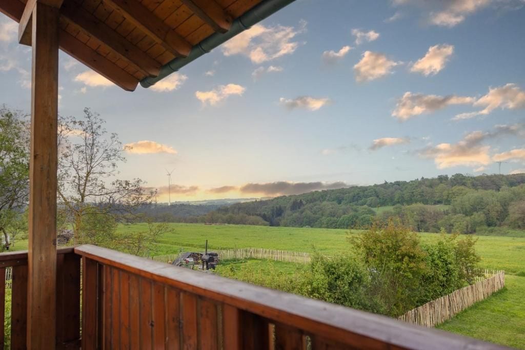A wooden deck overlooking a green field with a fence and trees, under a partly cloudy sky.