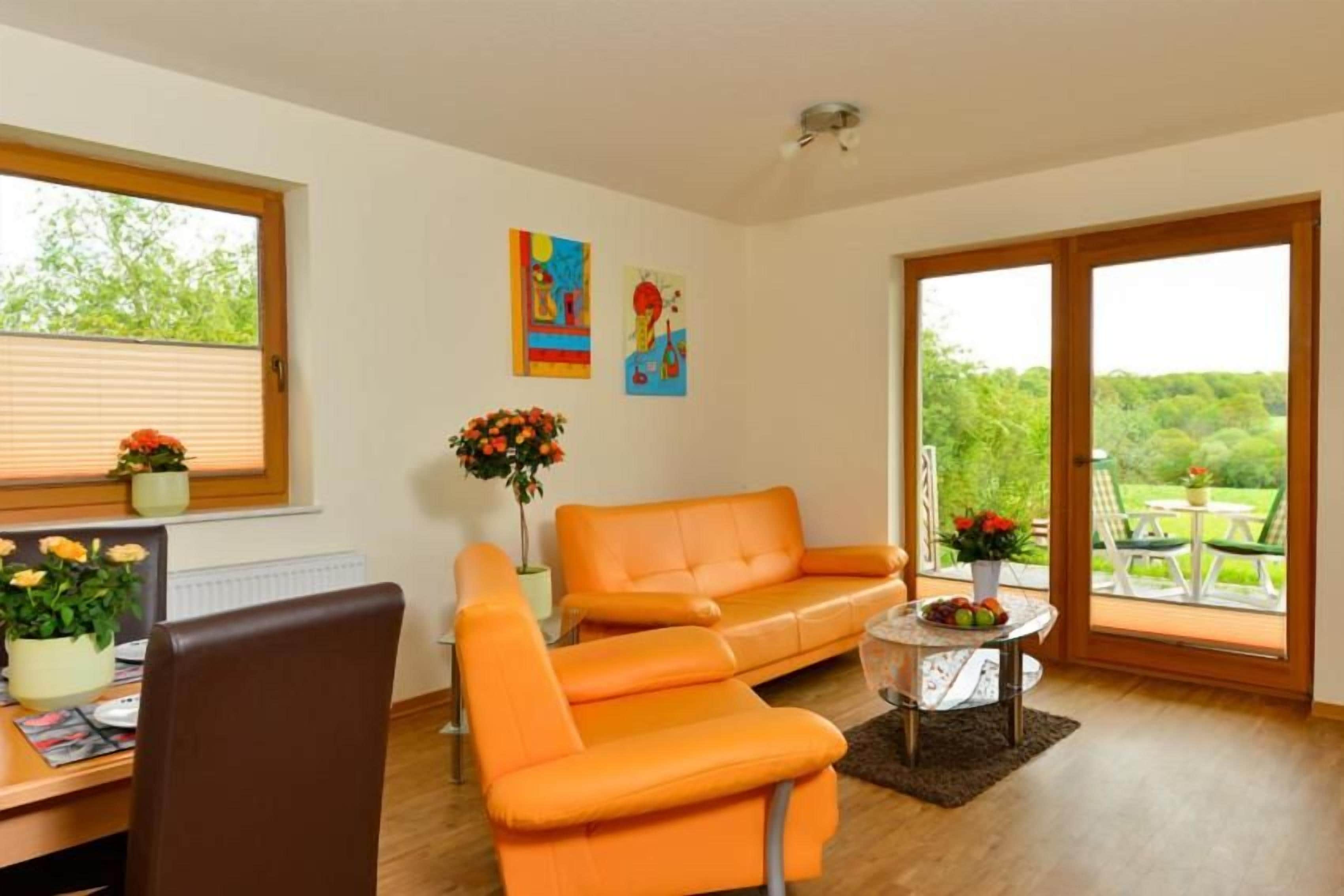 Une pièce avec un plancher en bois, une chaise marron, un canapé orange, une petite table ronde sur laquelle est posé un vase de fleurs, et une fenêtre avec des stores.