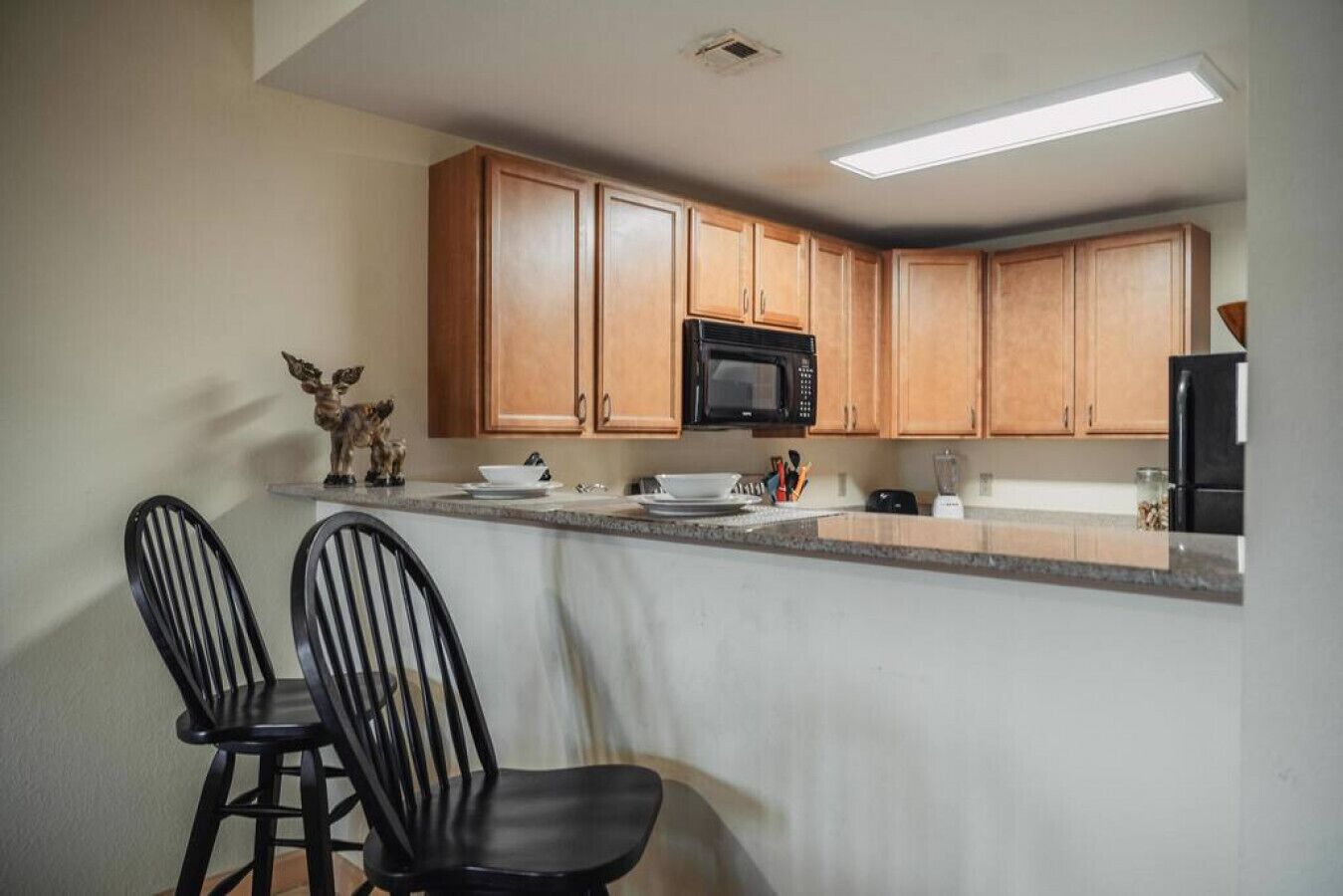 A kitchen with wooden cabinets, a microwave, a refrigerator, and two black chairs.