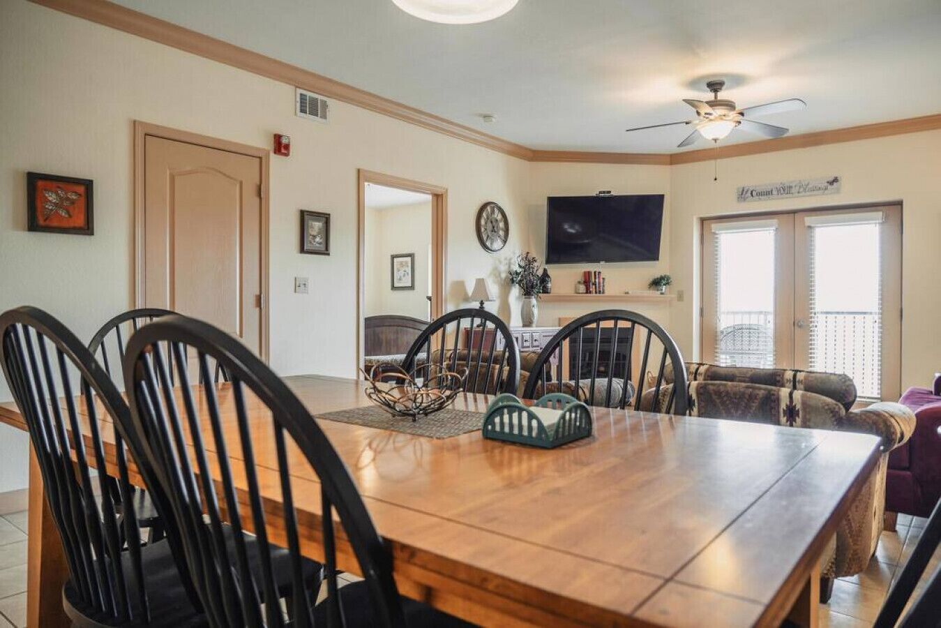 A dining area with a wooden table and chairs, a television mounted on the wall, and a sign that reads "Count Your Blessings".