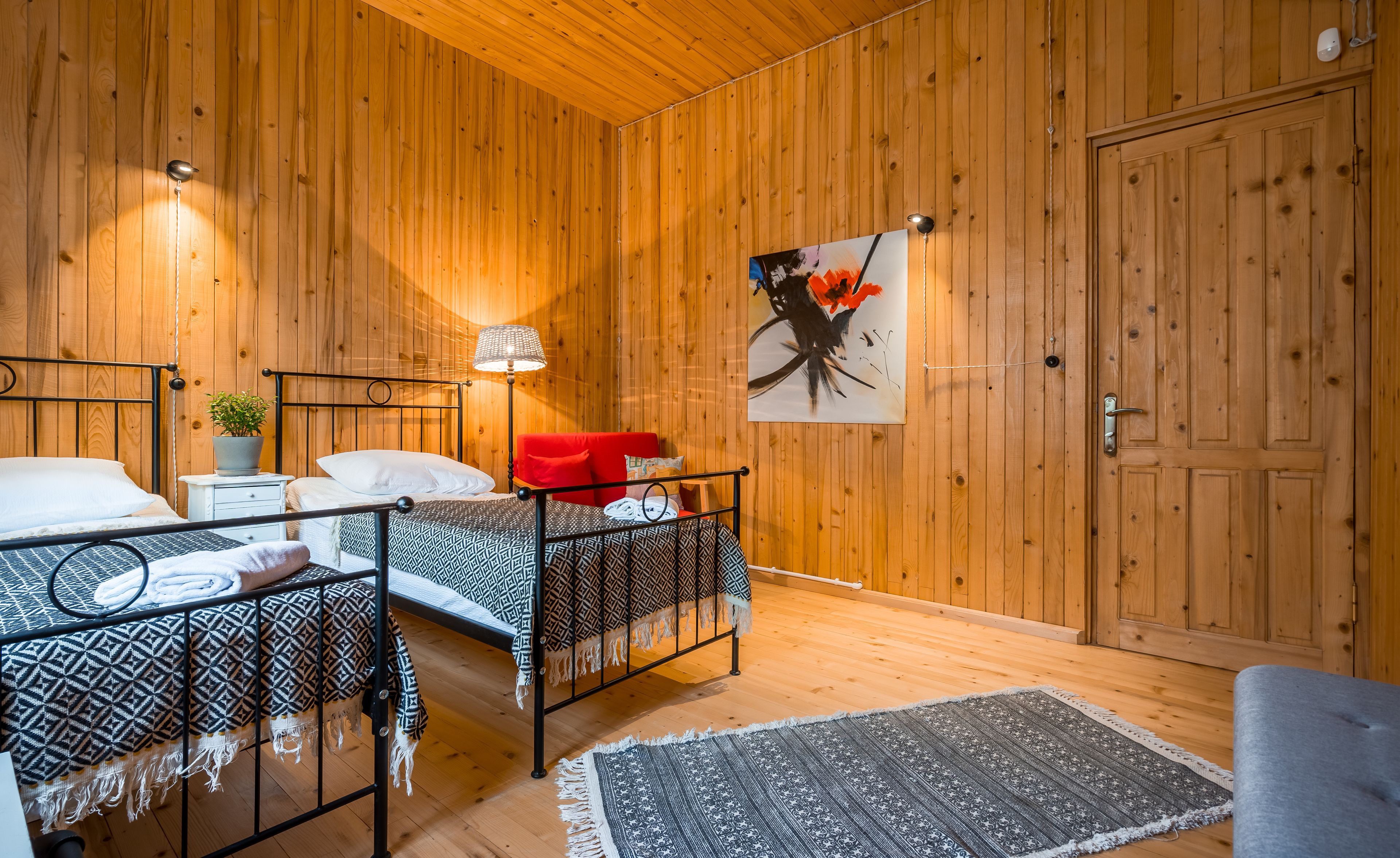 A wooden-paneled room with two beds, a red chair, a lamp, and a painting.