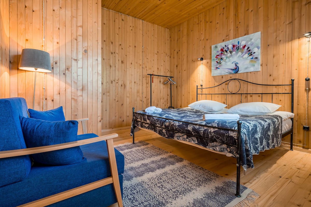 Una habitació amb panells de fusta, un llit, un sofà blau, un estotge amb dibuixos i una làmpada penjada a la paret.