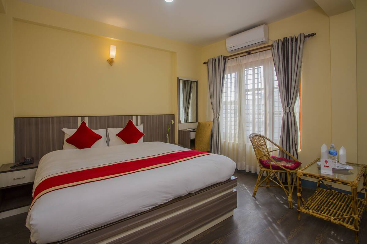 A hotel room with a large bed, a bedside table, a chair, a small table with a bottle, and an air conditioning unit.