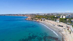 An aerial view of a coastal city with a wide beach, clear blue waters, and buildings along the shore.