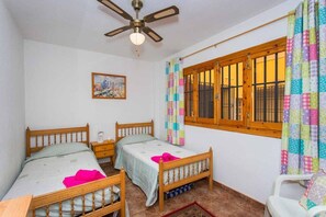 A bedroom with two wooden beds, a ceiling fan, a window with bars, and a colorful curtain.