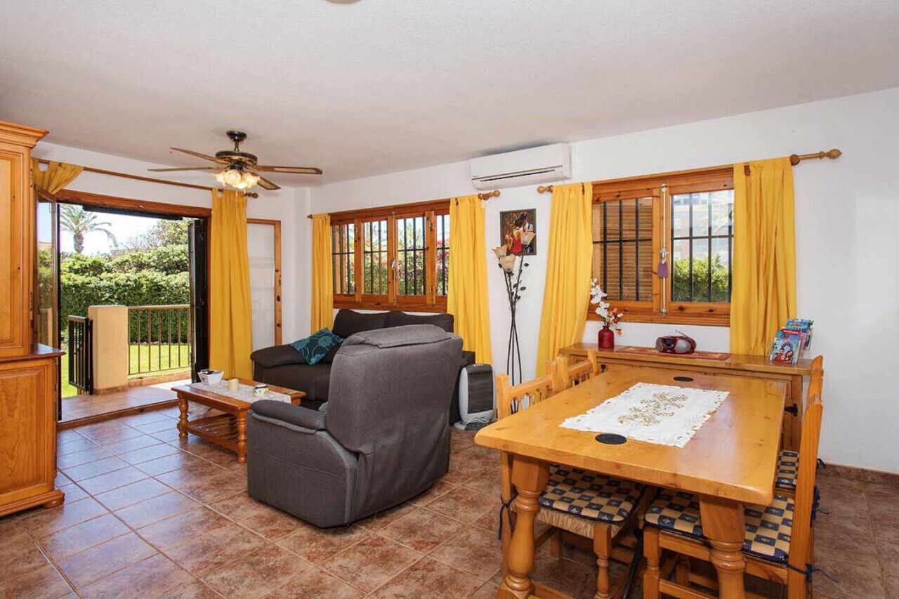 Uma sala de estar com sofá, mesa de jantar e cadeiras, ventilador de teto e grandes janelas com cortinas.
