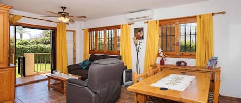 A living room with a sofa, a dining table with chairs, a ceiling fan, and large windows with curtains.
