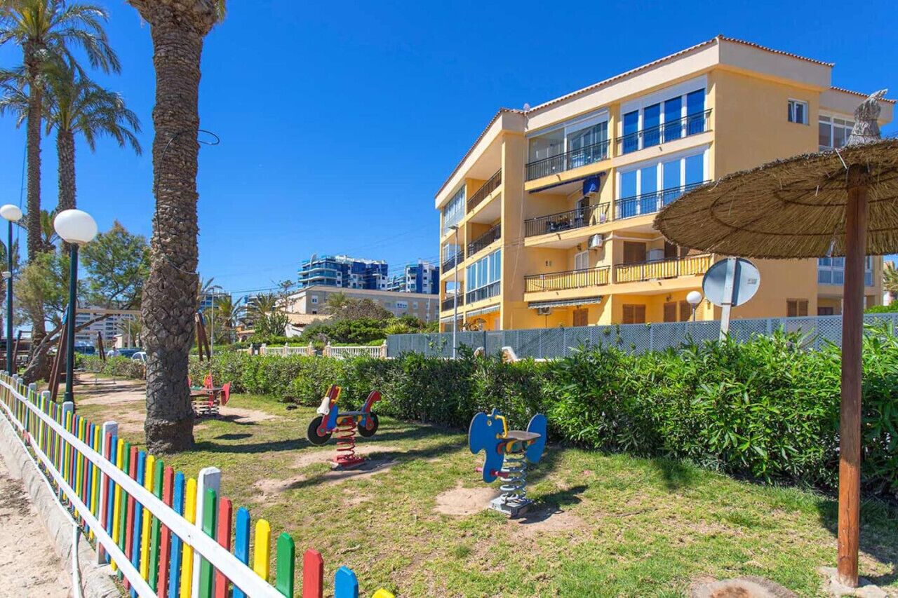 Un edificio de varios pisos con balcones, un patio de juegos cercado con juegos infantiles y palmeras.