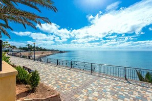 Un paseo marítimo con un sendero de baldosas, barandilla de metal y vista despejada al mar.