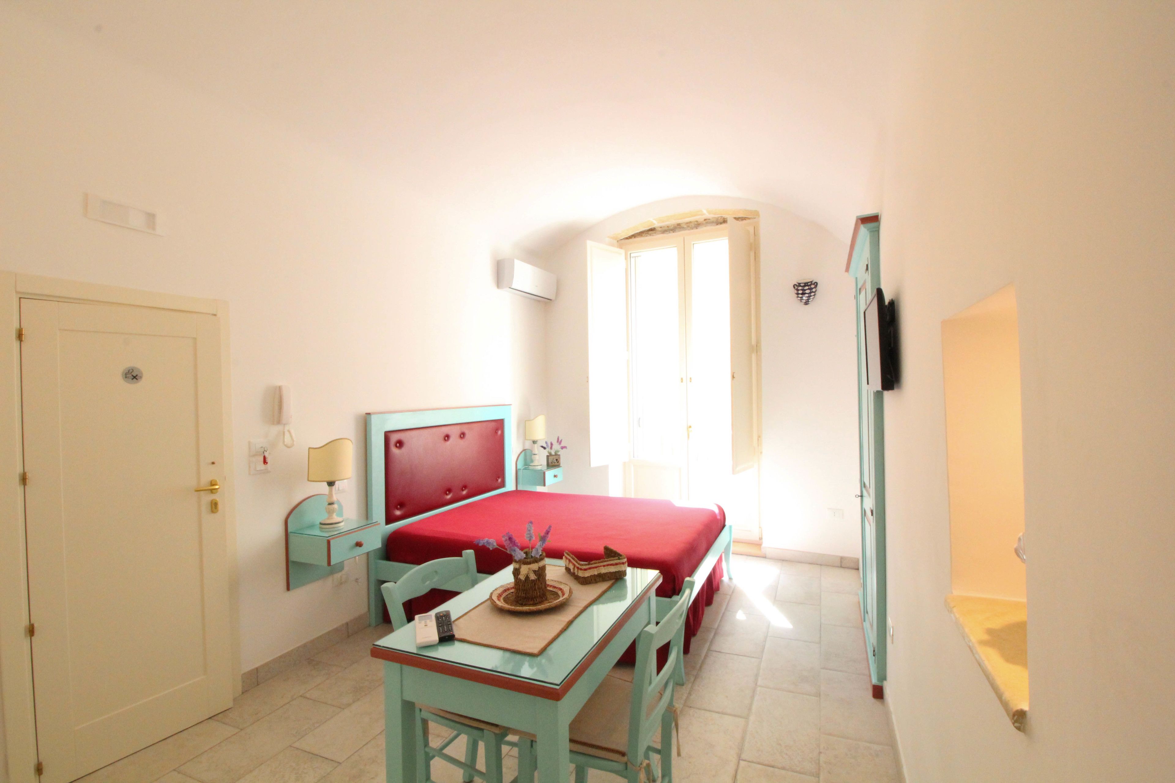 A room with a bed, a desk with a chair, a small table with a lamp, and a door.