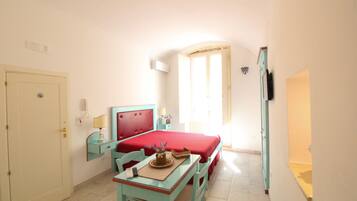 A room with a bed, a desk with a chair, a small table with a lamp, and a door.