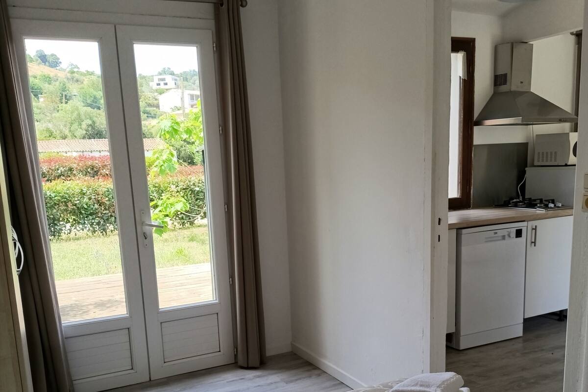 A hotel room with a bed, a view of the garden through a sliding door, and a kitchenette in the adjacent room.