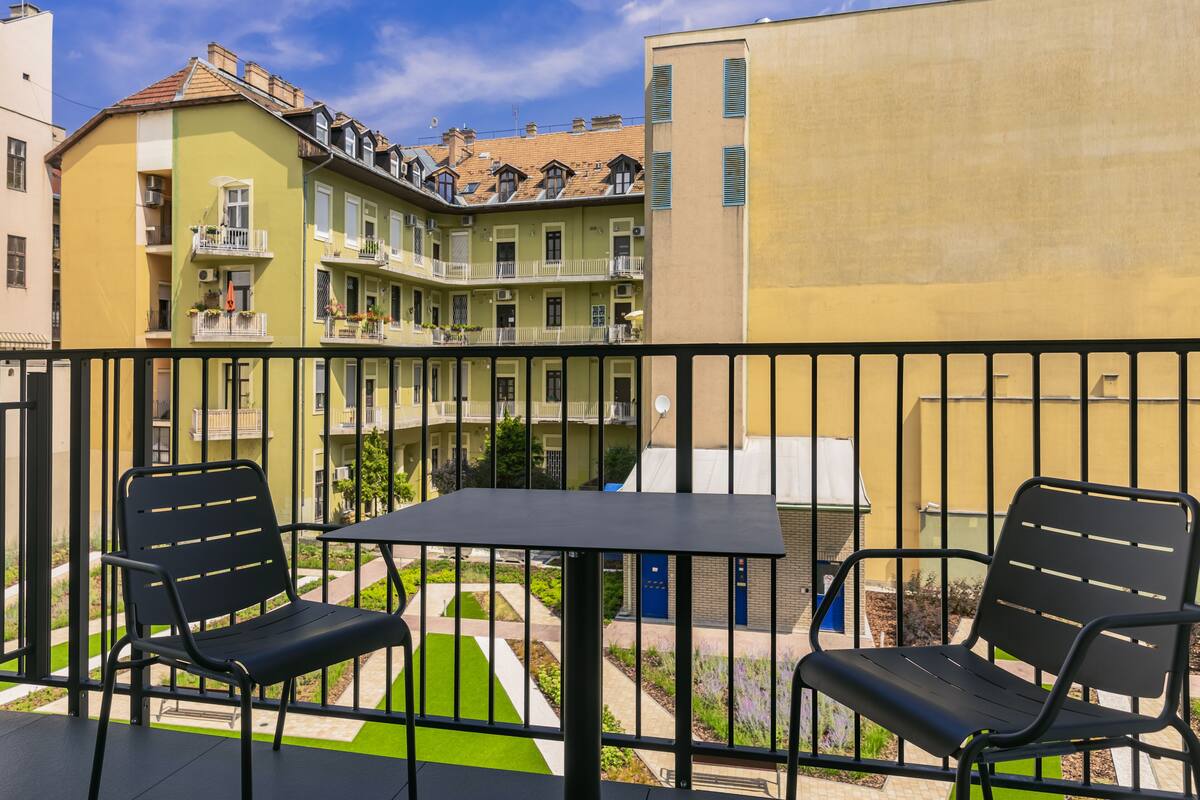 Un balcon avec deux chaises noires et une petite table, donnant sur une cour intérieure avec des bâtiments en arrière-plan.