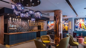 Zona de bar moderna con suelo de diseño geométrico, barra con rótulos 'Barcelona Budapest' y asientos coloridos.