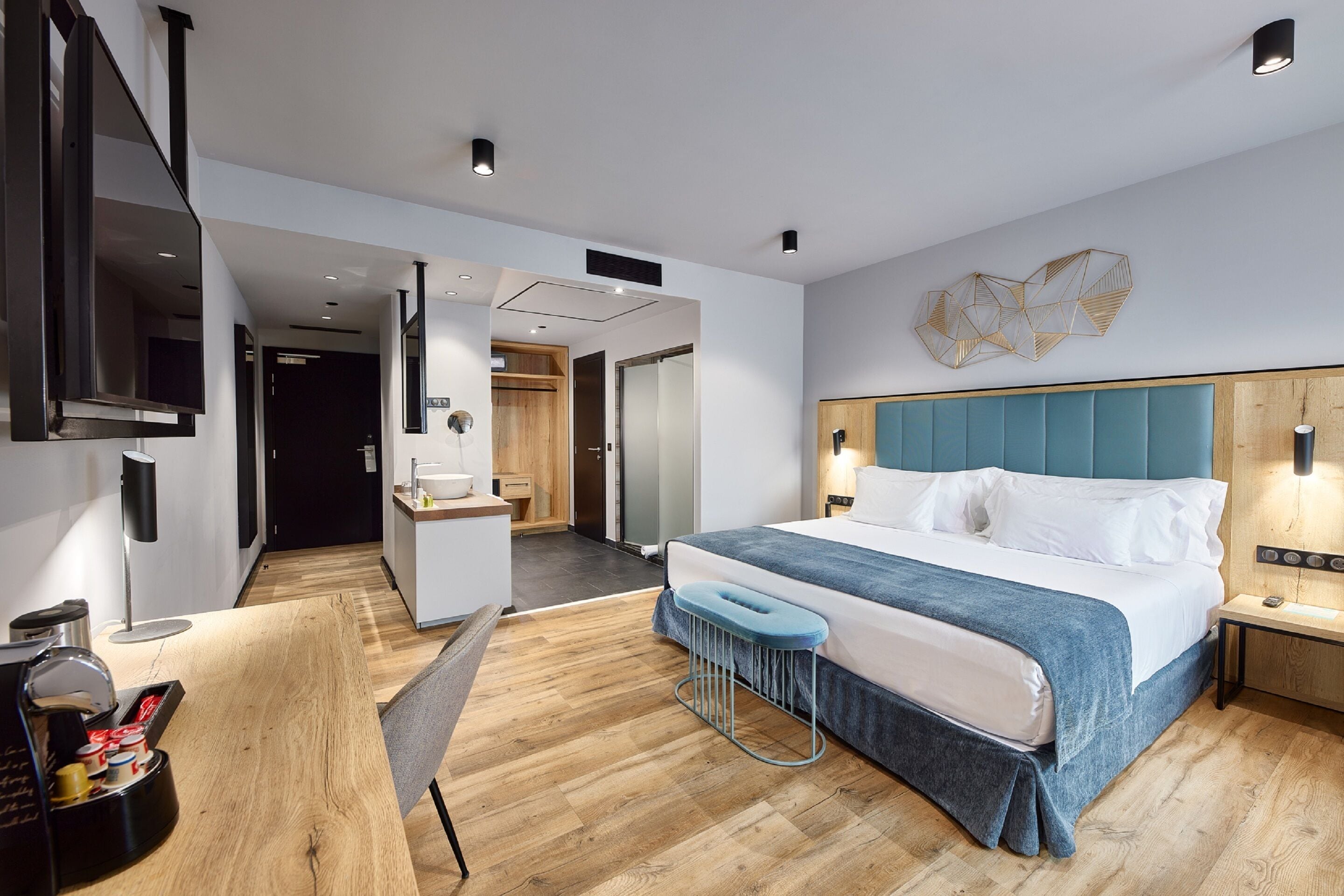 A modern hotel room with a large bed, a wooden floor, a desk with a chair, a small stool, and a TV mounted on the wall.