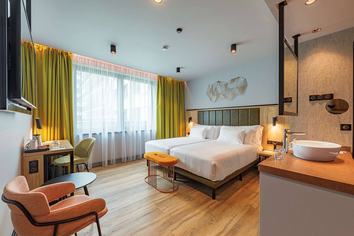 Une chambre d’hôtel avec un lit, une table de chevet, une lampe et une grande fenêtre avec des rideaux.