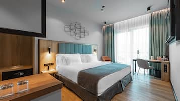 Quarto de hotel moderno com uma cama grande, uma escrivaninha e uma cadeira. A escrivaninha de madeira tem um computador e uma cadeira. O quarto possui uma janela com cortinas e uma televisão na parede.