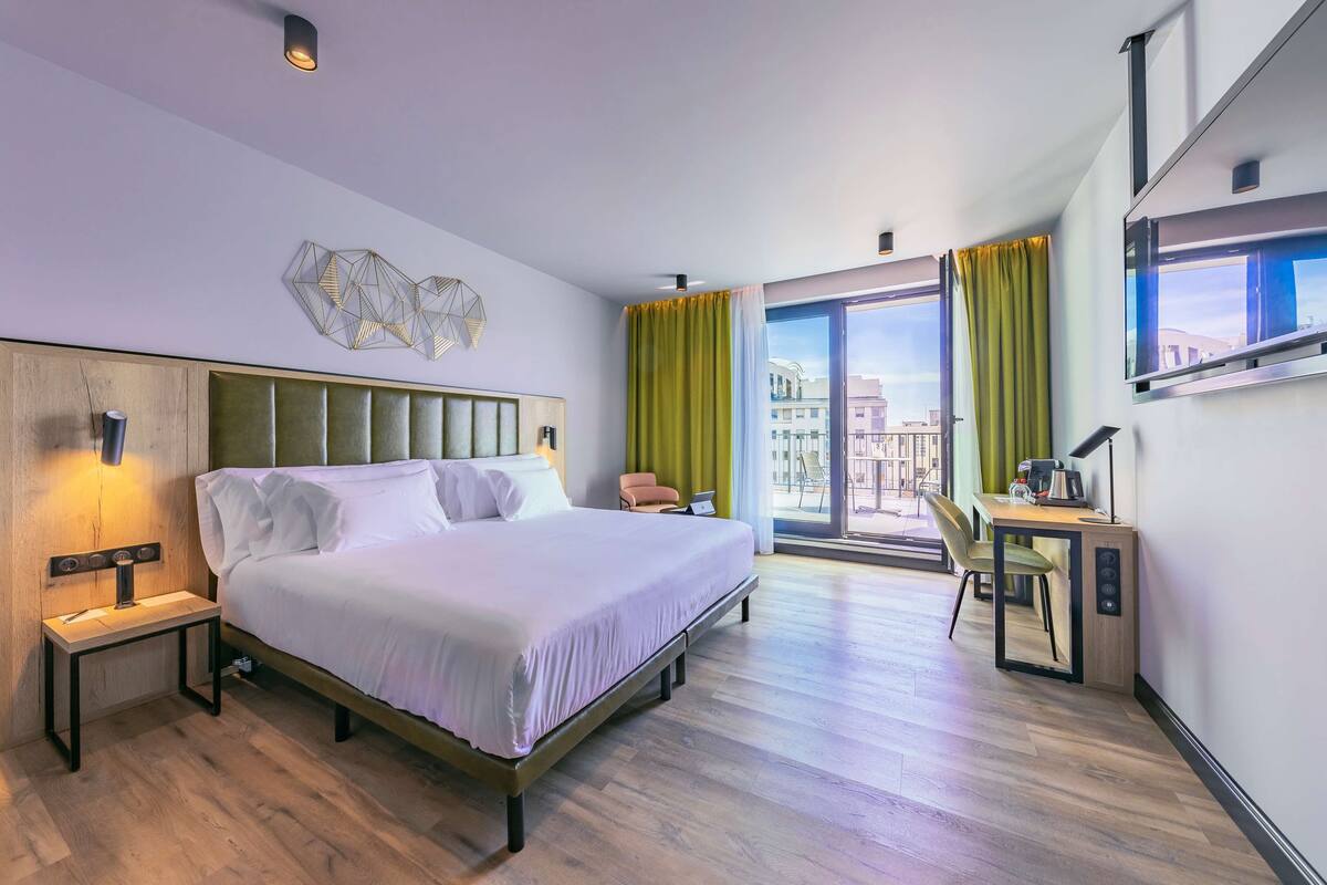 Une chambre d’hôtel moderne, dotée d’un grand lit, d’un bureau et offrant une vue sur la ville par la fenêtre.