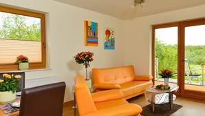 A modern living room with a sofa, a chair, a small table with a vase of flowers, and a window with blinds. - Ferienwohnung Panoramablick (Kastellaun)
