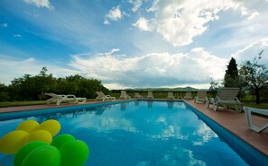 Una piscina con sillas de descanso y globos, rodeada de follaje y colinas bajo un cielo nublado.