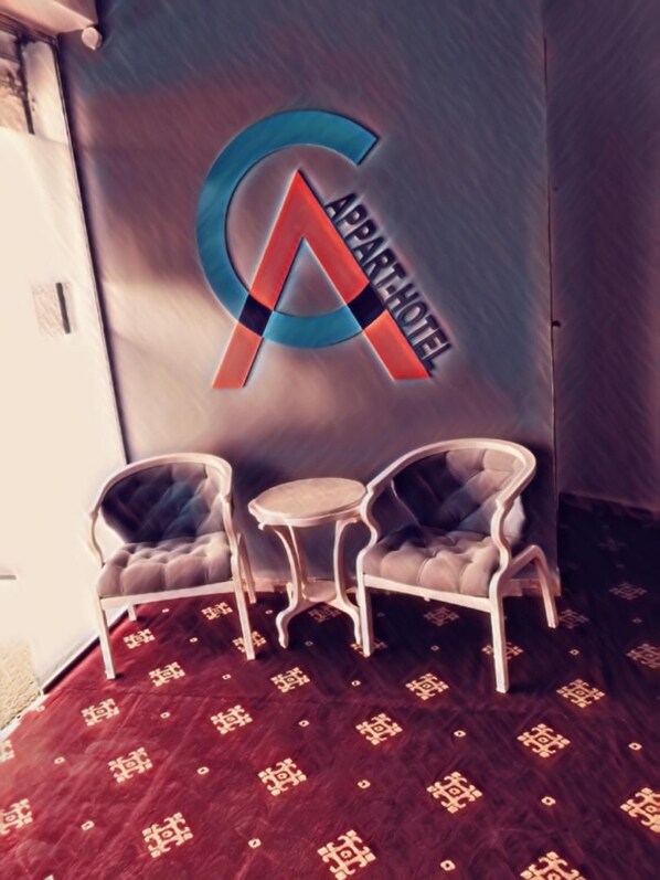 A hotel room with a large logo on the wall, two upholstered chairs, a small round table, and a patterned carpet.