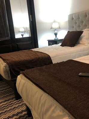 A hotel room with two beds, each with a brown blanket, a bedside table with a lamp, and a window with curtains.
