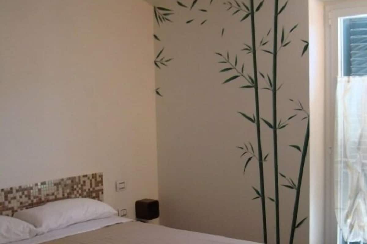 A bedroom with a bed, a wall with bamboo decals, a window with a curtain, and a small nightstand with a lamp.