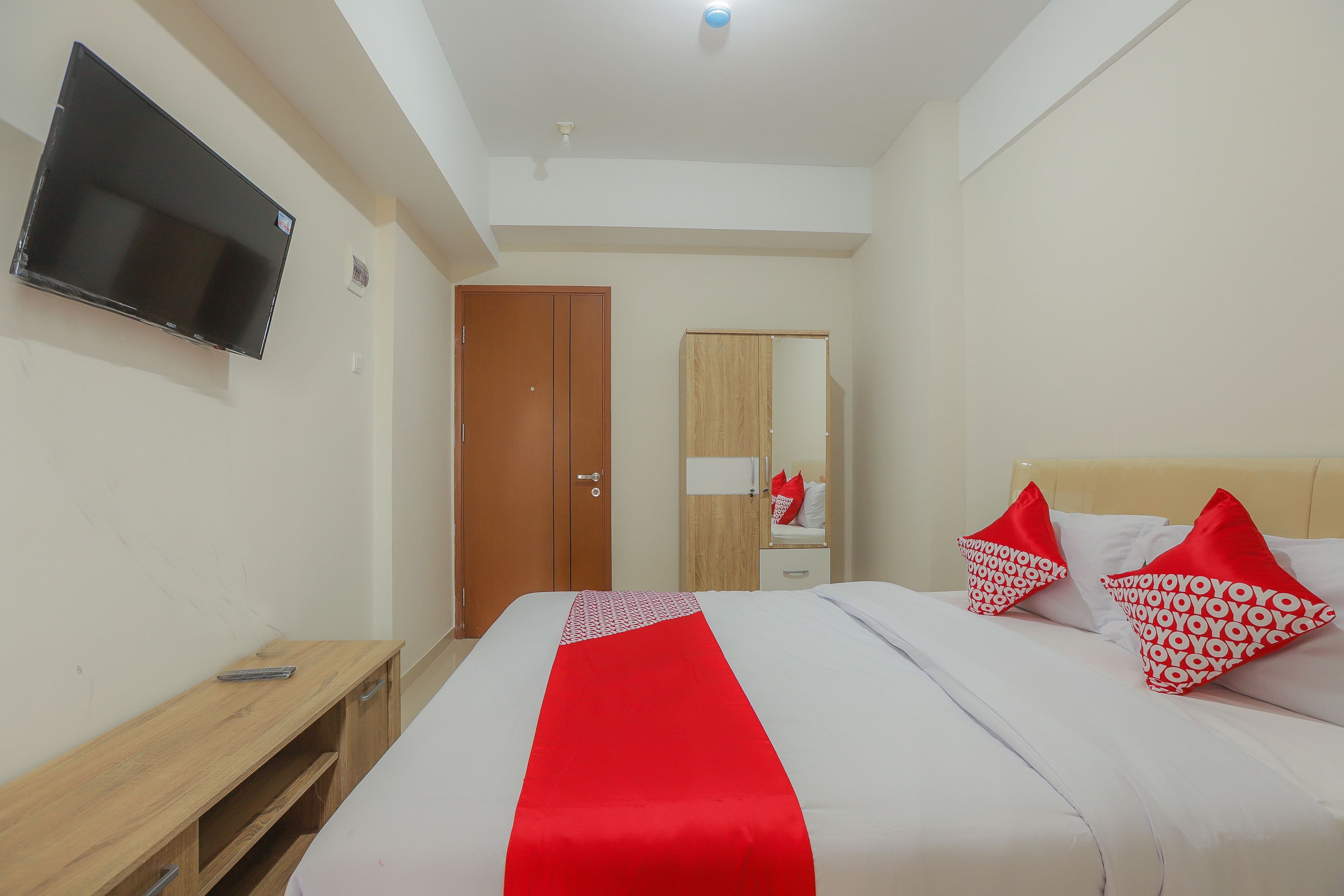 A hotel room with a bed, a TV mounted on the wall, a wooden nightstand, a wardrobe, and a mirror.