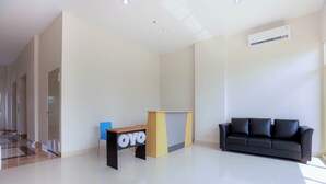 A spacious, modern room with a black leather sofa, a wooden desk with a blue chair, and a large window providing natural light. - OYO 630 Green Lake View Cimanggis (Depok)