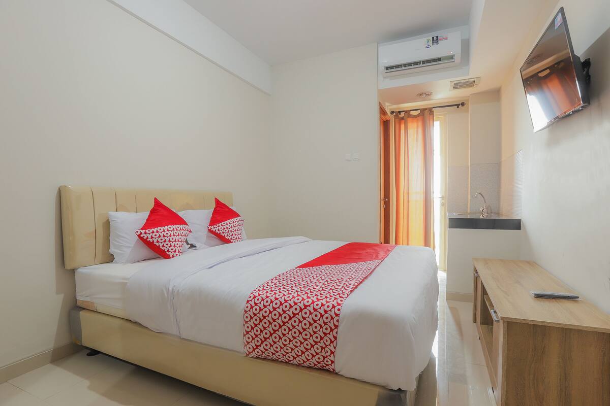 A neatly made bed with a white and red patterned bedspread, a wooden bedside table with a remote control, and a wall-mounted TV.