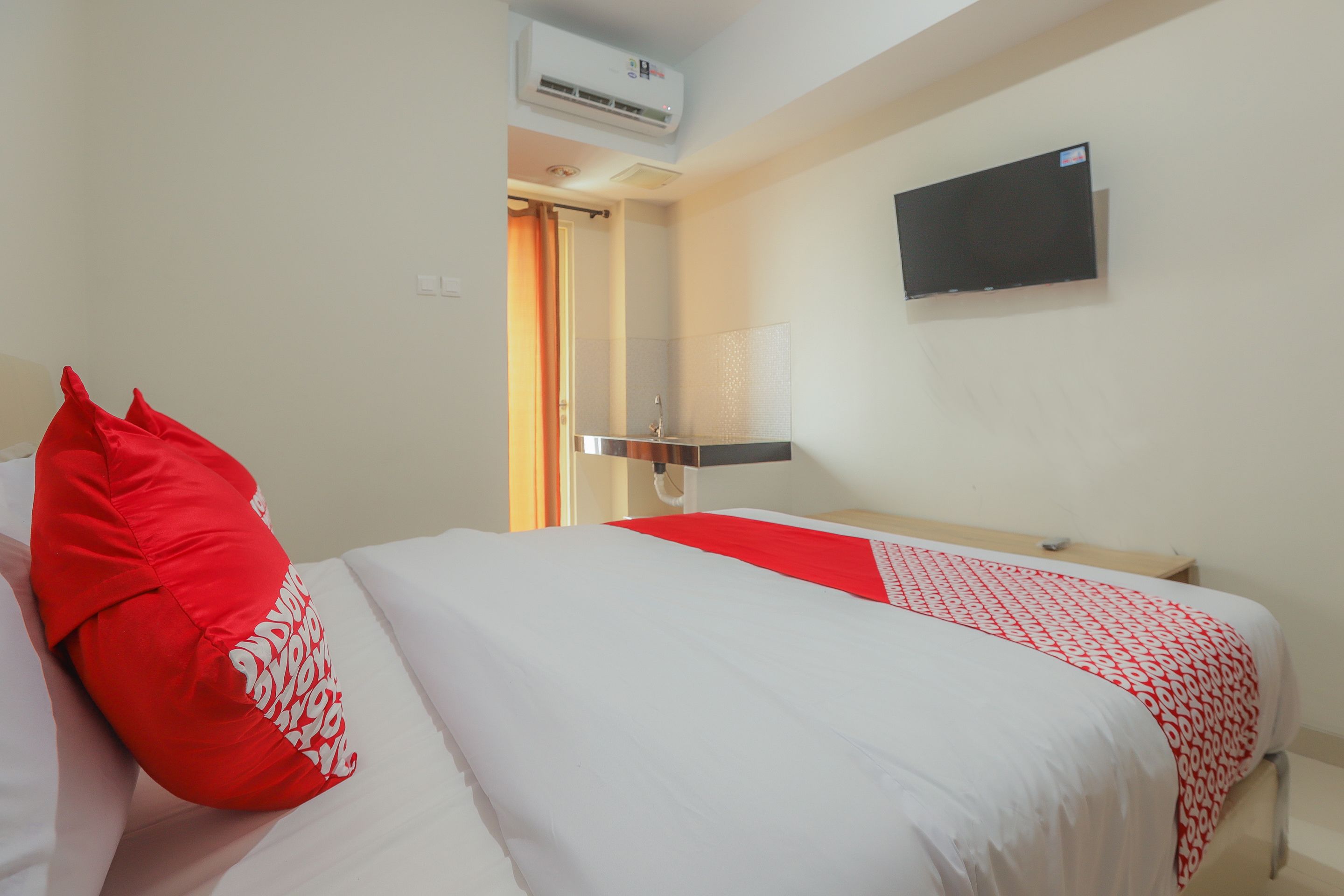 Une chambre d’hôtel avec un lit, un oreiller rouge, un couvre-lit blanc, un chemin de lit rouge et blanc à motifs, un téléviseur fixé au mur et un appareil de climatisation.