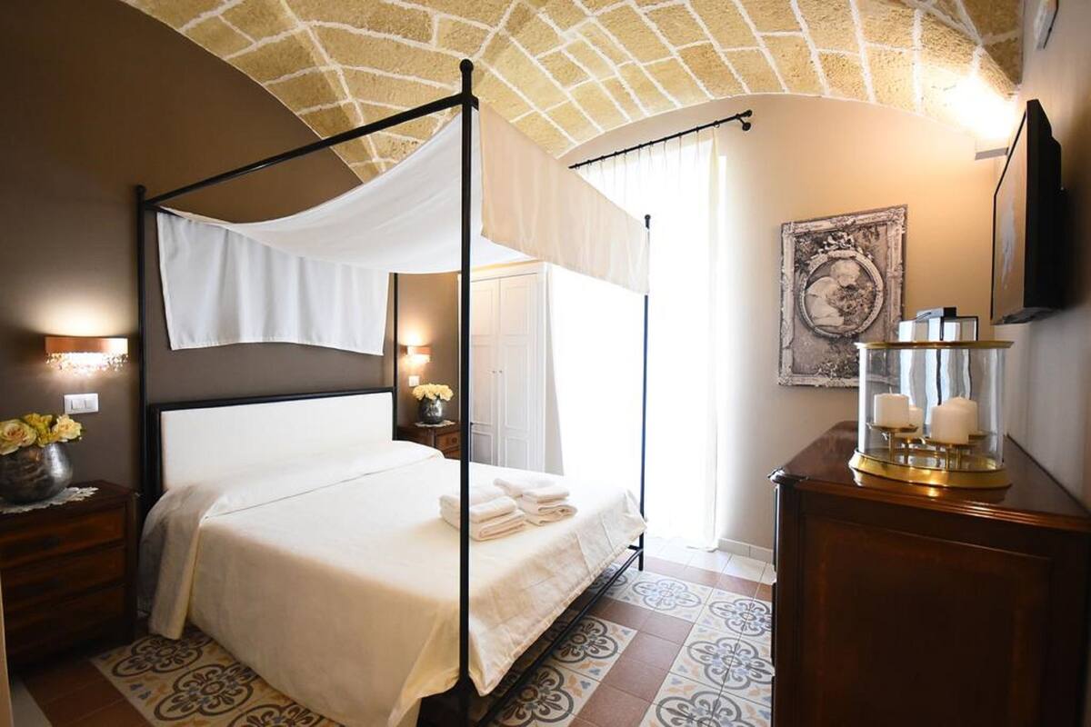 Una cama con dosel con ropa de cama blanca, una mesita de noche con un florero y un televisor montado en la pared, además de un candelabro de vidrio decorativo.