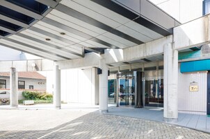 Entrada de un edificio moderno con un vestíbulo cubierto, puerta giratoria de vidrio y toldo azul.
