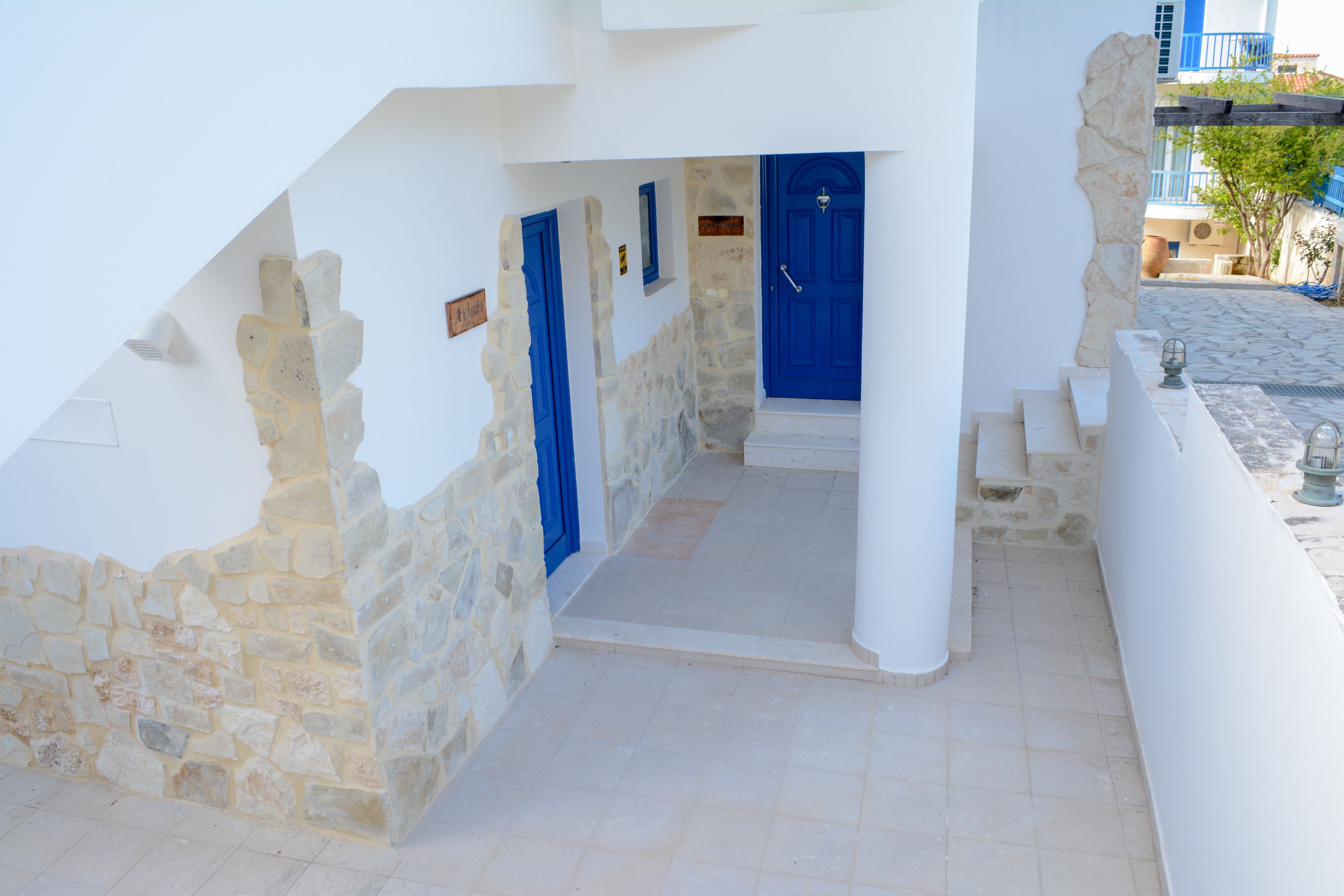Un estrecho sendero de piedra conduce a un conjunto de puertas azules, flanqueado por paredes blancas y una pared de piedra en un lado.
