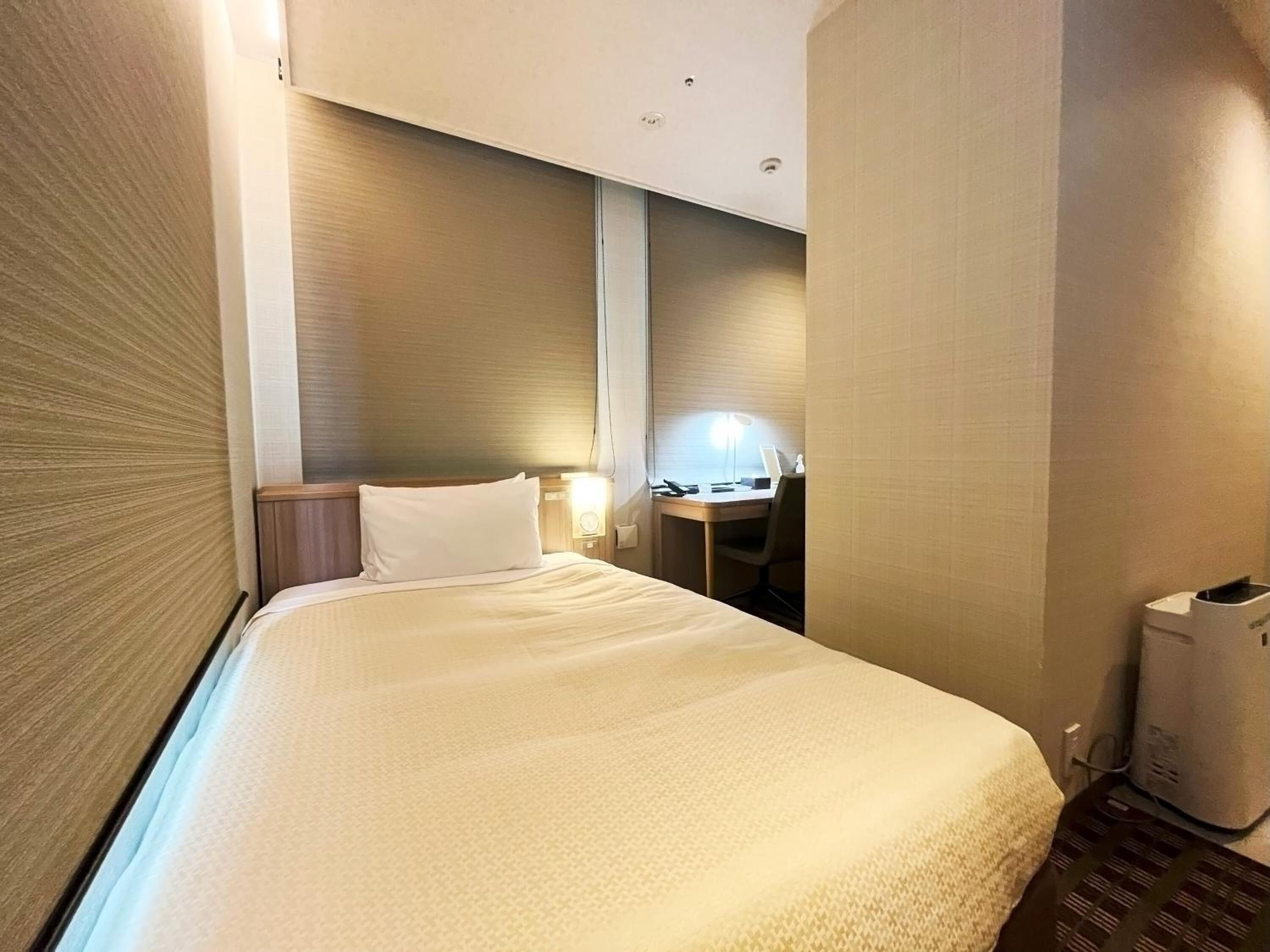 A hotel room with a bed, a desk, a chair, and a lamp.