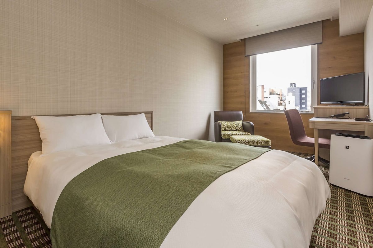 Une chambre d’hôtel avec un lit, un bureau, une chaise et une grande fenêtre.