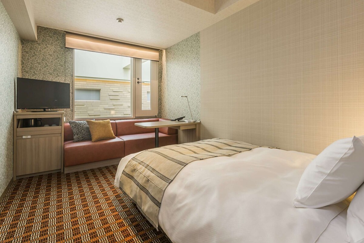 A hotel room with a bed, a TV on a stand, a seating area with a table, and a patterned floor.