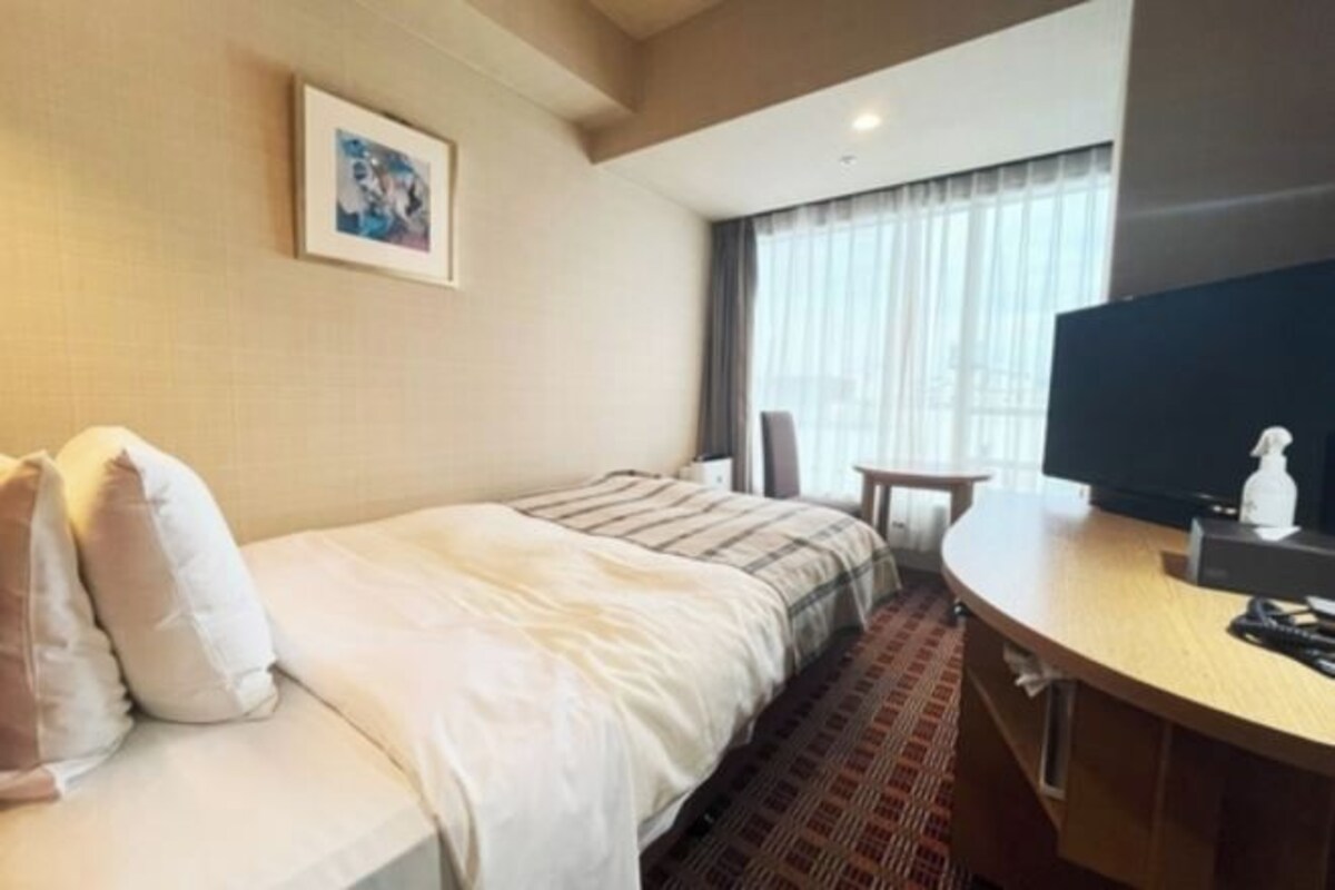 A hotel room with a bed, a TV, a small table, and a window with curtains.