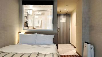 A modern hotel room with a large bed, a window with a view of the bathroom, and a small heater on the floor.