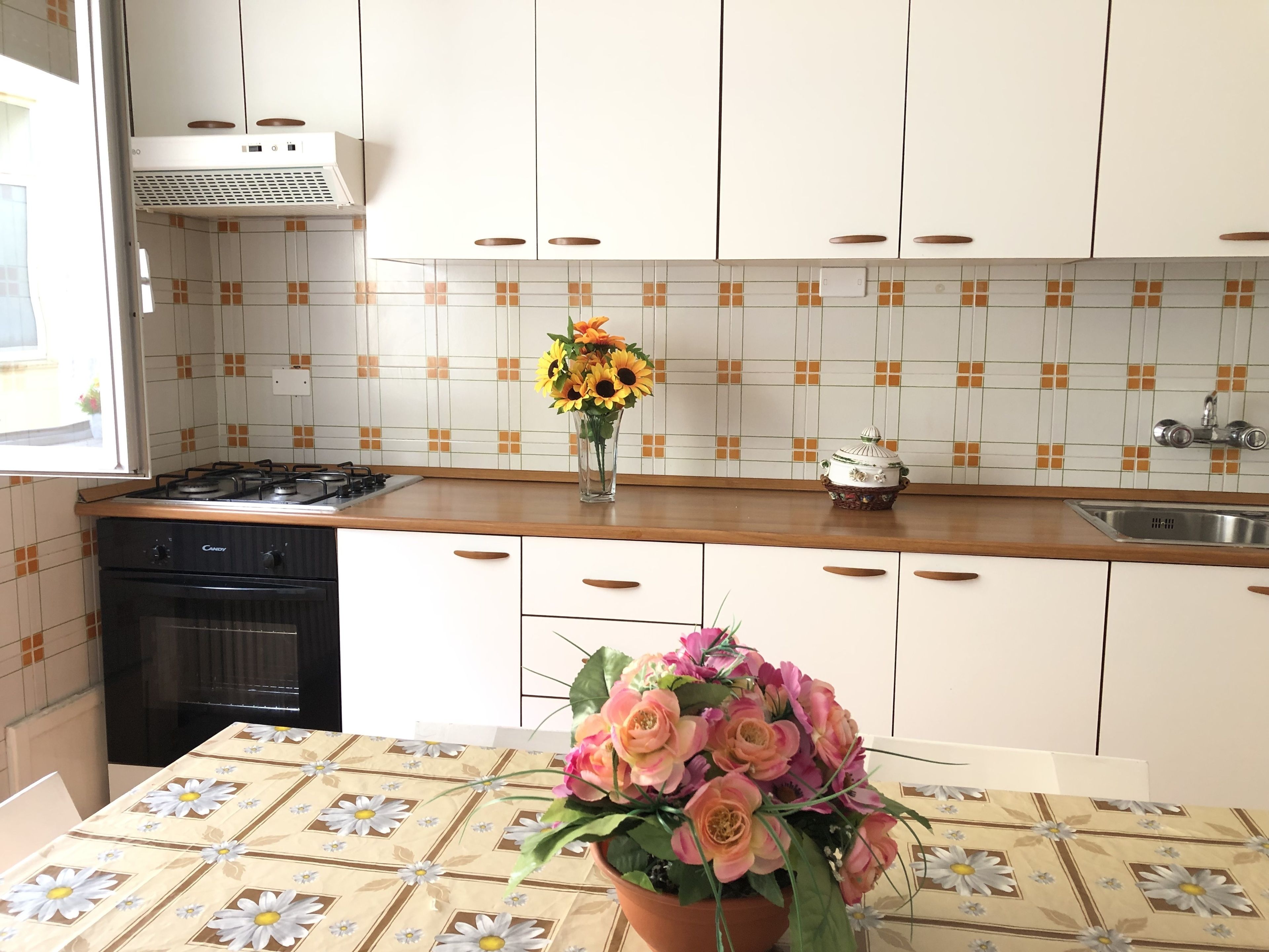 Une cuisine avec des meubles blancs, une cuisinière, un évier et une table recouverte d’une nappe à motifs floraux.