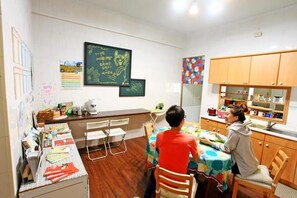 Huone, jossa on ruokapöytä, tuoleja, keittiöalue kaappeineen, tauluseinä, jossa on kirjoitusta, sekä erilaisia esineitä seinillä.