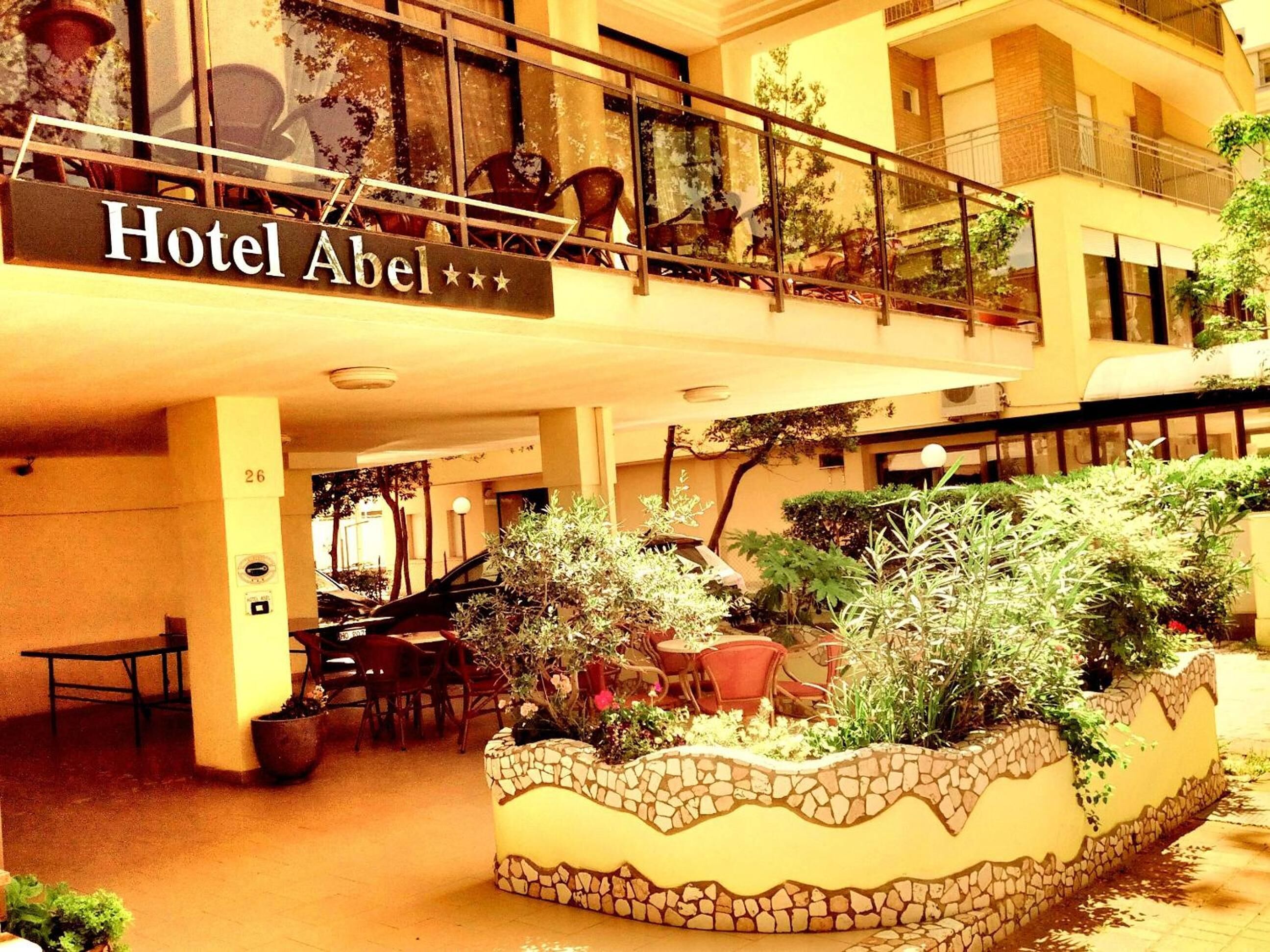 Un interno di hotel con un cartello che indica 'Hotel Abel'. Sono presenti aree salotto all'aperto con tavoli e sedie, e un vaso decorativo con piante.