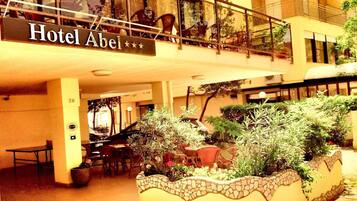 Un interno di hotel con un cartello che indica 'Hotel Abel'. Sono presenti aree salotto all'aperto con tavoli e sedie, e un vaso decorativo con piante.