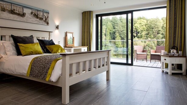 A bedroom with a bed, a bedside table, a chair, and a balcony with a view of trees. - Canada Lodge and Lake (Cardiff)