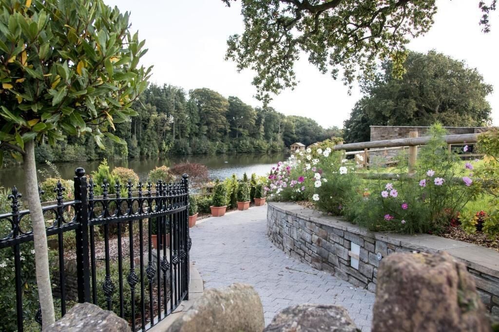 A paved pathway leading to a body of water, surrounded by greenery and a stone wall.